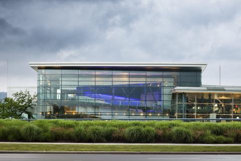 Exterior view of a square building with glass walls and a white roof. Green bushes, a sidewalk, and grass are in the foreground.