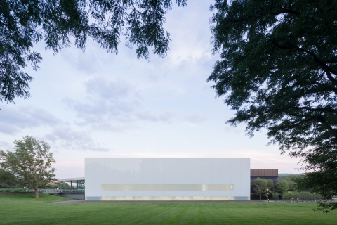Exterior view of a square building with glass walls and a white roof. Green bushes, a sidewalk, and grass are in the foreground.