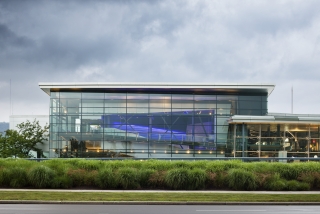 Exterior view of a square building with glass walls and a white roof. Green bushes, a sidewalk, and grass are in the foreground.
