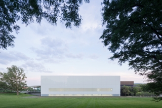 Exterior view of a square building with glass walls and a white roof. Green bushes, a sidewalk, and grass are in the foreground.