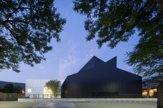 White square building next to a black, angular building at dusk, with green trees in the foreground.