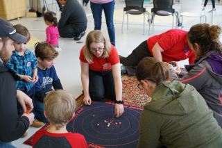 A group of visitors and volunteers sit next to a mat with marbles on it.