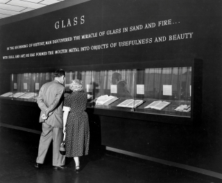 Black and white photo of two people standing looking through a narrow glass case at books on display in a museum. The image is from the 1950s.