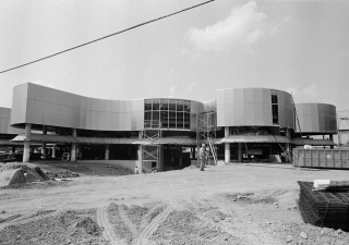 Black and white photo of a metal clad building on concrete pillars under construction