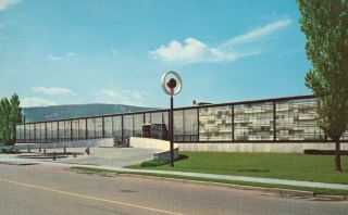 1951 photograph of the Corning Museum of Glass. A glass, rectangular building with a lawn, trees, and a glory-hole-inspired logo on a pole are in front of the building.